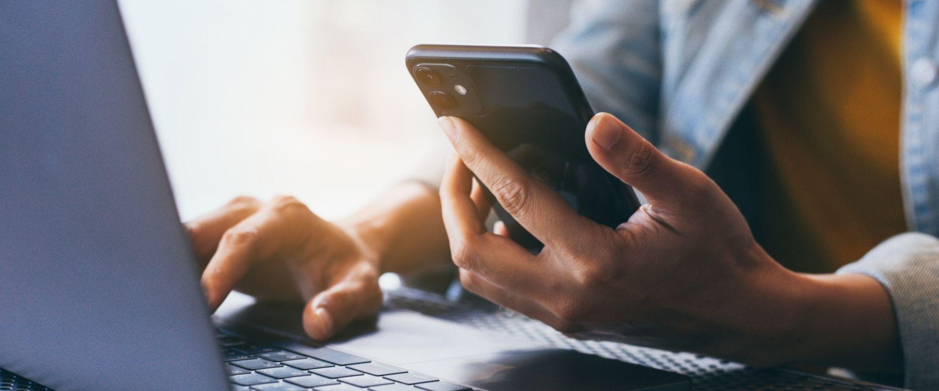 female sitting at laptop typing and holding a cell phone